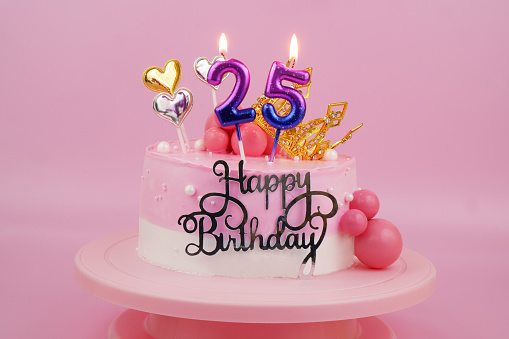 Pink birthday cake with gold crown and burning candles, numbers 25 on pink background.