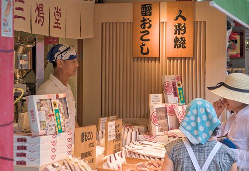 A seller of baked sweets in Nakamise Street, serving two female customers who are tasting the local specialty. Tokyo, Japan, 21_8_2019.