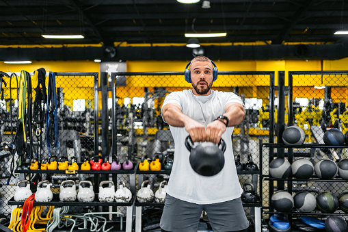 Handsome young muscular man training in the gym with a kettlebell while listening to music via headphones in the Kuwait City.