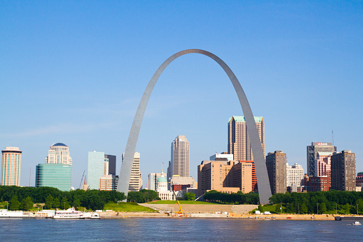 Iconic Gateway Arch dominates St. Louis skyline against a backdrop of blue sky. Vibrant cityscape with high-rise buildings and leisure boats on the river. Perfect for architecture, tourism, and city branding.