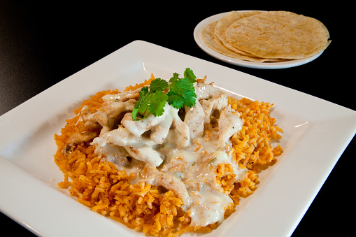 Delicious Mexican Arroz con Pollo with Creamy Queso Sauce and Vibrant Mexican Rice under layer of chicken on a Square White Plate. Perfect for Restaurants, Cookbooks, and Food Blogs. Tortillas on side plate