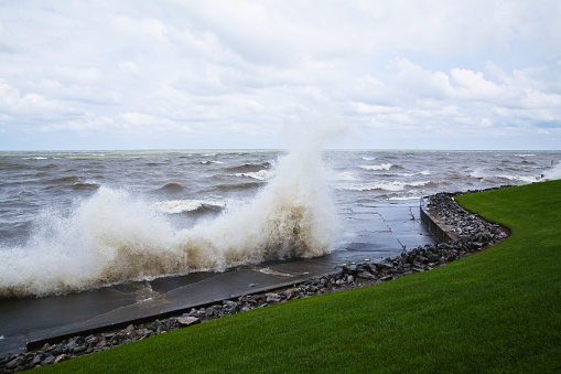 Dynamic clash of nature and structure as dark waves crash against seawall in overcast weather, creating a striking contrast with serene green lawn. Perfect for illustrating environmental challenges and resilience.