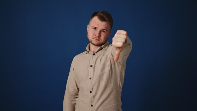 Man in his 30s on blue background giving thumbs up then thumbs down, changing opinion