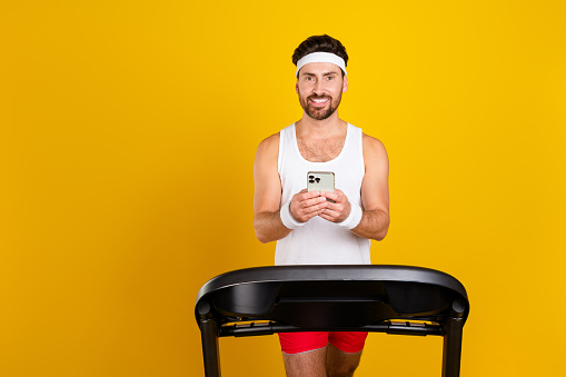 Photo of cool cheerful guy activewear walking treadmill texting apple iphone samsung gadget empty space isolated yellow color background