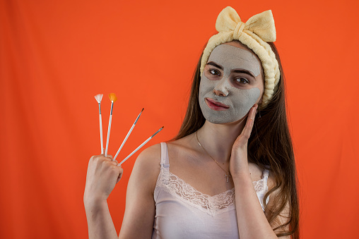 Young caucasian woman applying with brush green clay mask to her face isolated on orange. Beauty treatment for perfect skin