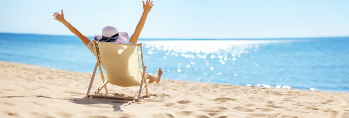 Happy woman sitting with raised hands in the chaise-lounge on the sea beach. Tourism and vacation concept.