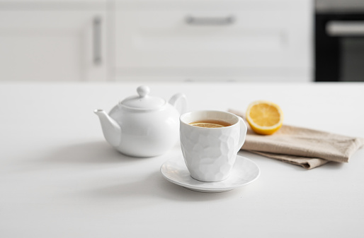 A cup of tea with lemon and a teapot on a white table against the background of a white kitchen. Interior in modern Scandinavian style. Breakfast concept. Copy space.