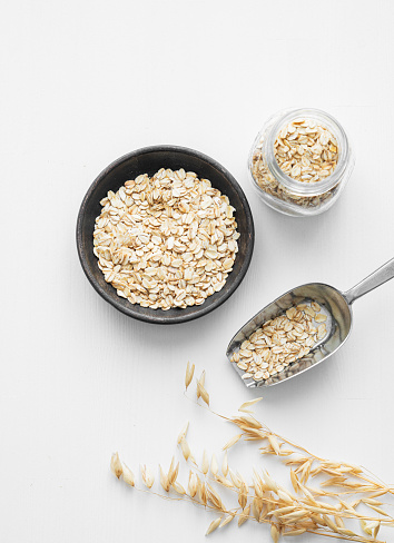 Oatmeal in a plate, jar and in a scoop on a white textured background with dry branch. The concept of a natural vegan cereal product for preparing a healthy breakfast. Top view and copy space.