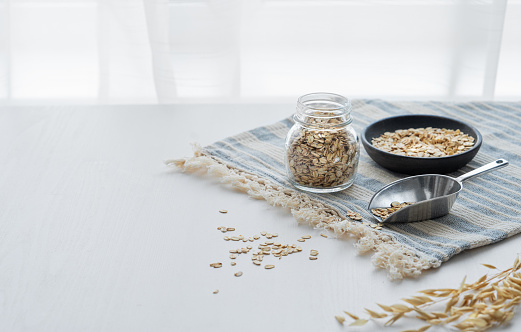 Oatmeal in a jar and in a bowl on a napkin on a white textured table in front of the kitchen window. The concept of a natural vegan cereal product for preparing a healthy breakfast. Copy space.
