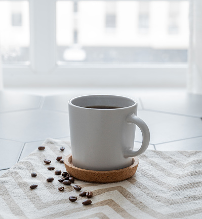 A cup of black coffee on a napkin on a trendy gray tile background with coffee beans and morning light from the window. Breakfast drink concept. Copy space.