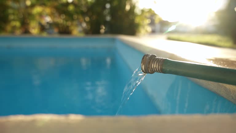 Close up stream of water flowing from a green hose and filling a clean blue swimming pool in the backyard at sunset. Weekly pool maintenance and cleaning