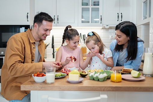 Young Caucasian family, parents with daughters making healthy breakfast at the modern kitchen