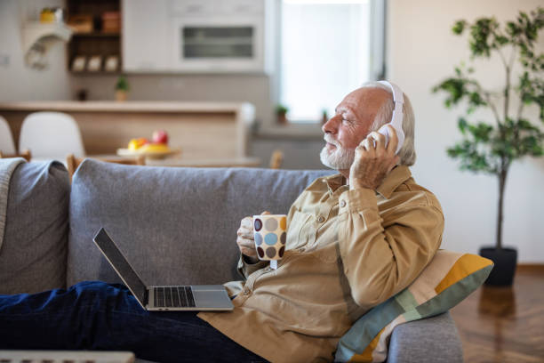istock Happy elderly man at home using laptop. Happy elderly man relaxing on a sofa at home and listening to music on laptop with headphones, while drinking coffee. 2010255424