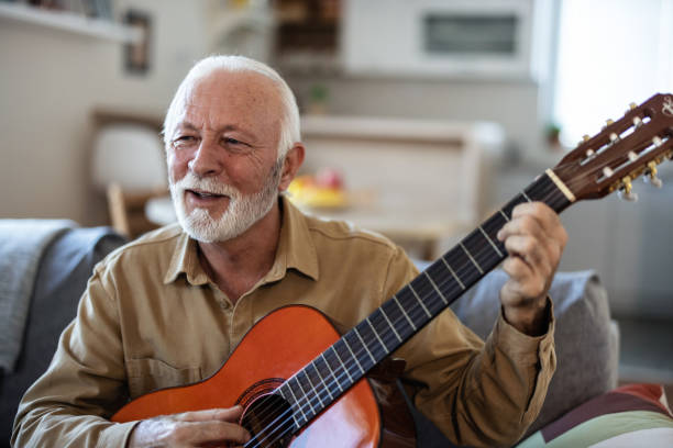 istock Bearded senior man with acoustic instrumental guitar. Man playing guitar while sitting on sofa in light living room. 2010265605