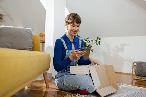 Happy, young woman sitting at home and opening a box with a just delivered order.