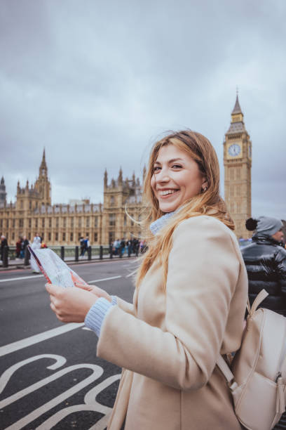 istock Young white tourist girl in her 30s smiling and looking in camera while holding a printed city map near Big Ben clock tower  in London, United Kingdom - creative stock image 2010286551