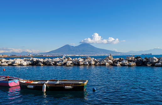 A serene scene of Naples, Italy, with a majestic Vesuvius volcano looming in the background, a bustling marina filled with docked boats, and a clear blue sky dotted with fluffy clouds