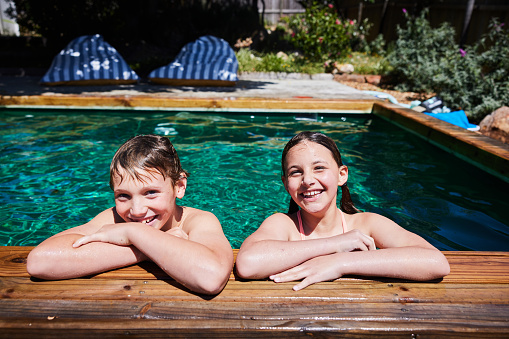 Portrait of a smiling young brother and sister swimming together in their swimming pool at home in summer