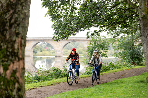 LGBTQI+ lesbian couples a bike ride together in a public park in Hexham, North East England. They are cycling side by side along a rivers edge.