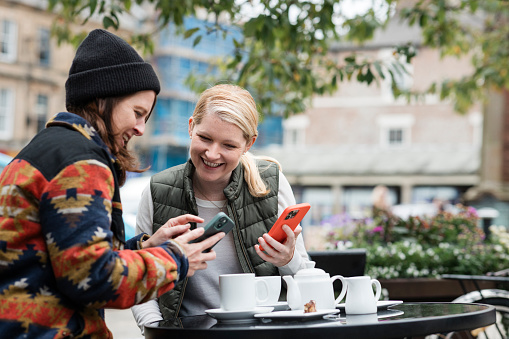 LGBTQI+ lesbian couple sitting outdoors at a cafe together enjoying a cup of tea. They are looking at their mobile phones laughing at them.