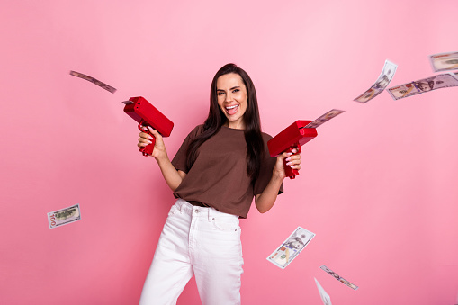 Portrait of nice positive lady toothy smile hands hold money gun shoot dollar bills isolated on pink color background.