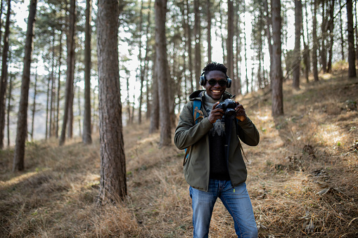 Young smiling black freelance photographer looking at the camera and smiling. He is holding his digital camera. Millennial male tourist, hiker love to photographing wilderness area.