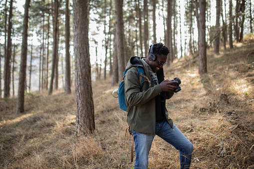 Millennial black man, freelance photographer, with headphones is hiking through the forest, listening music and photographing . He is resting and looking at the photos he made with his camera.