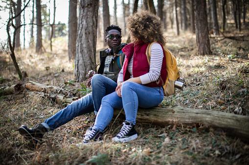 Millennial couple hiking through the forest and enjoying in beautiful nature. Young black man and young white woman with curly hair now taking a break and they are sitting on the log, relaxing and talking to each other.