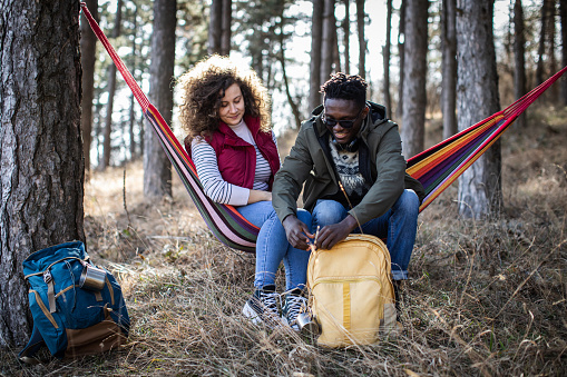 Adventurous millennial couple are sitting and relaxing in a hammock. They are taking a break from hiking through the forest. Enjoying in beautiful nature. Active lifestyle.