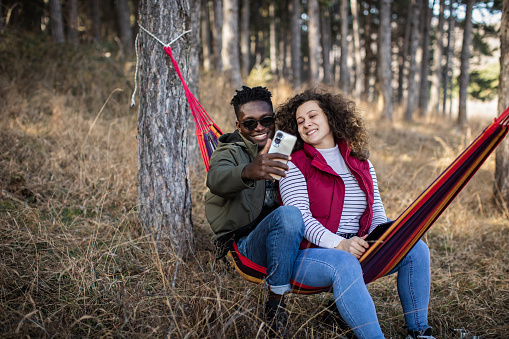 Adventurous millennial couple are sitting and relaxing in a hammock. They are taking a break from hiking through the forest. Young black man with sunglasses and young white woman with curly hair are smiling and making a selfie.