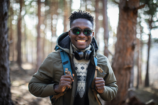 Young beautiful black man with backpack hiking through the forest.  He is wearing sweater and jacket. He is posing, smiling and looking at the camera. He has a sunglasses, a headphones around his neck and he crossed his arms. Trees is all around him.