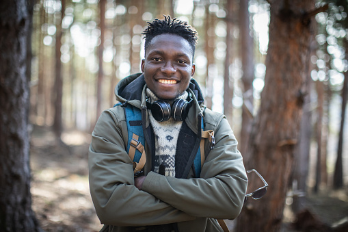 Young beautiful black man with backpack hiking through the forest.  He is wearing sweater and jacket. He is posing, smiling and looking at the camera. He has a headphones around his neck and he crossed his arms and holds sunglasses in his hand. Trees are all around him.