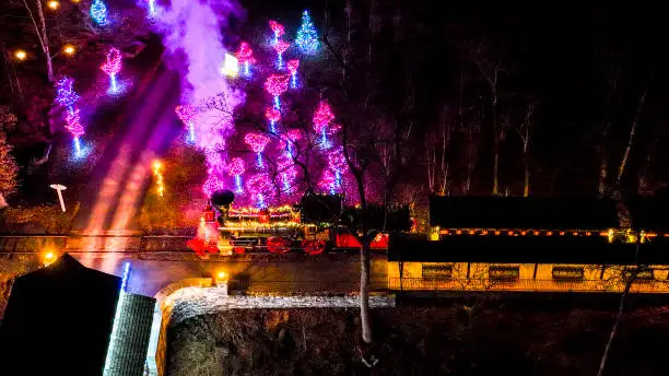 Nighttime Aerial Shot Of An Illuminated Steam Locomotive With Red Wheels Releasing Purple Smoke Next To Decorated Trees.