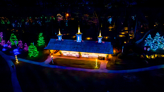 Overhead Night View Of A Building With Illuminated Rooflines And Steeples Surrounded By Trees Decorated With Bright, Colorful Lights.