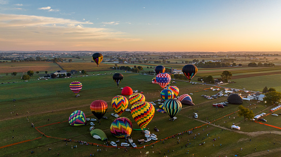 An Aerial View of Cluster Of Colorful Hot Air Balloons Preparing For Flight At Dawn With Spectators And Vehicles Gathered On A Rural Field.