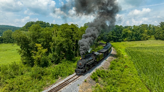 An Aerial View of a Classic Black Narrow Gauge Steam Train Chugging Along Tracks Surrounded By Lush Greenery And Emitting Thick Smoke Into The Clear Sky.