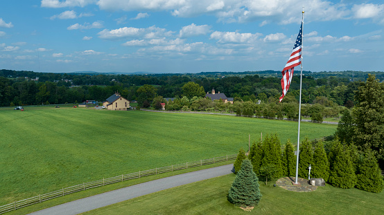 Expansive Green Field Edged By A Winding Path And Wooden Fences, With A Prominent American Flag Flying High, Overlooking A Rural Landscape Dotted With Buildings And Trees Under Scattered Clouds.