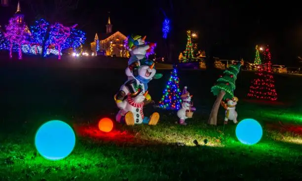 Nighttime Holiday Scene With Inflatable Snowman Figures And Brightly Lit Orbs On The Ground, Set Against A Backdrop Of Trees And Bushes Adorned With Colorful Lights.