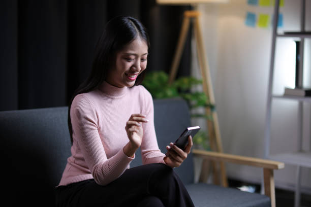 istock Young Asian woman sitting on sofa at home using mobile phone to send text messages and talk on the phone with her friends. 2010419801