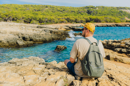 Rear view of a male in yellow cap enjoying weekend at the beautiful remote rocky beach with view of green pine forest in Gallipoli peninsula, Puglia region, South Italy