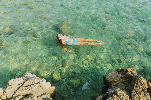 High-angle view of smiling female in blue swimsuit enjoying swim in crystal blue waters in Porto Selvaggio National Park, Puglia, South Italy