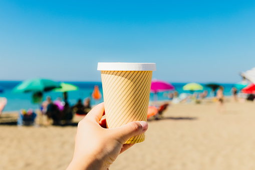 Close-up view of woman's hand holding paper cup with Cappuccino during morning at the yellow sand beach with sun umbrellas in Puglia, Italy