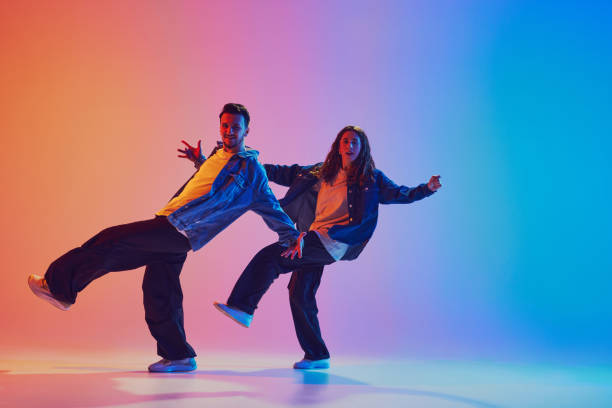 istock Dance duo mid-action in modern urban style outfits, raises one leg while dancing in motion, against gradient background in neon light. 2010497898