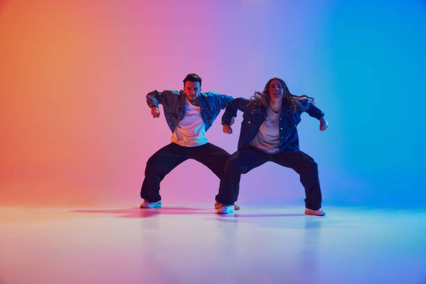 istock Energetic dance pair in denim jackets in squatting dance pose in neon-lit studio against gradient blue-orange background. 2010497901