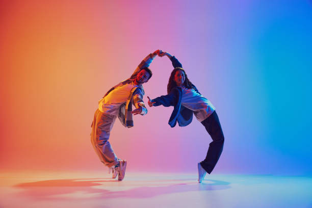 Dynamic shot of young dance duo holding hands while dancing in motion against gradient background in neon light.