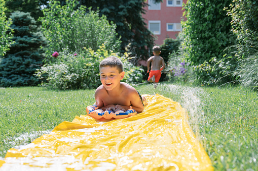 Happy summer scene of two boys sliding trough a yellow wet plastic in the garden