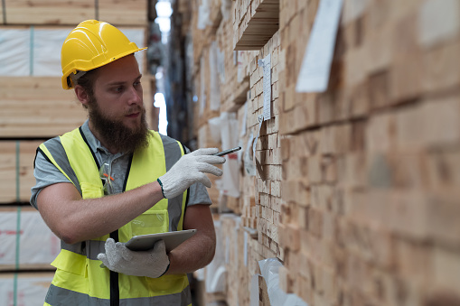Male warehouse worker using digital tablet checks stock inventory in lumber storage warehouse. Male worker checks barcodes on pile of plank wooden in wooden warehouse