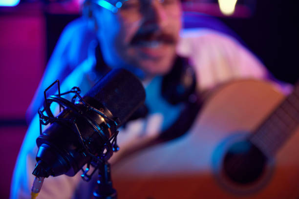 istock Studio microphone against blurred background with male artist, blogger, playing guitar and recording music in neon light. 2010540613