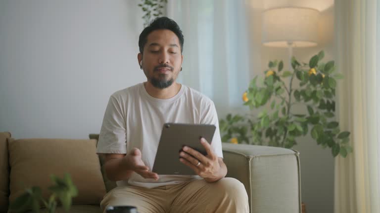 Middle-aged Asian male e-commerce businessman sitting on the sofa using a tablet in a meeting at home.