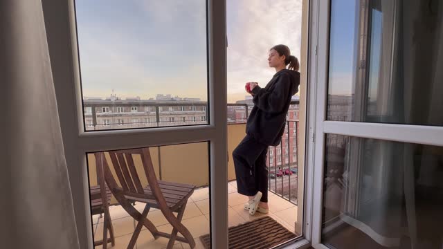 The girl stands on the balcony of the apartment and drinks tea looking at the city